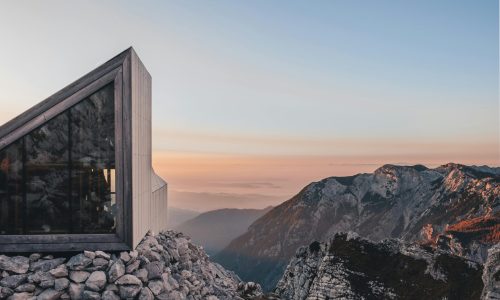 A sleek modern cabin perched on rocky terrains in Kranj, Slovenia, with stunning mountain views at sunset.