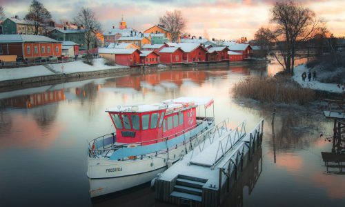 Snow-covered boats reflecting in Porvoo's historic harbor during a winter sunset.