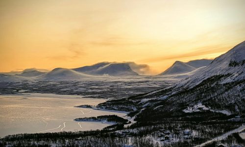 Breathtaking scenery of frozen lake surrounded by snowy mountains with leafless trees against sunset sky